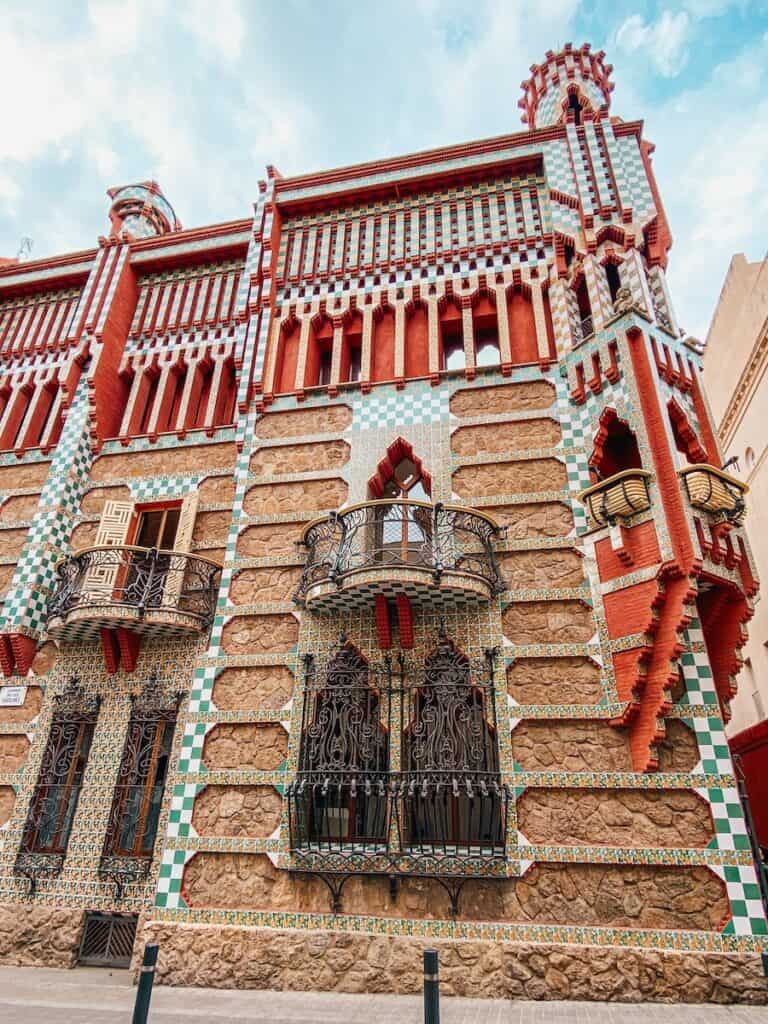 This image showcases Casa Vicens, one of Antoni Gaudí's early architectural works in Barcelona. The building features a distinctive façade with intricate wrought iron balconies, vibrant geometric tiles, and a mix of red brick and stone, highlighting Gaudí's innovative use of materials and color. The combination of Moorish and Catalan styles reflects his evolving architectural approach.