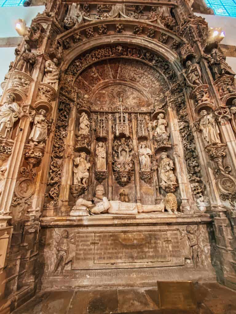 An intricately carved Gothic tomb with elaborate stonework and religious statues stands within a historic church. The reclining effigy of a knight rests on the sarcophagus, with a lion at his feet symbolizing courage. Above, delicate sculptural details and an arched canopy showcase the grandeur of medieval craftsmanship.