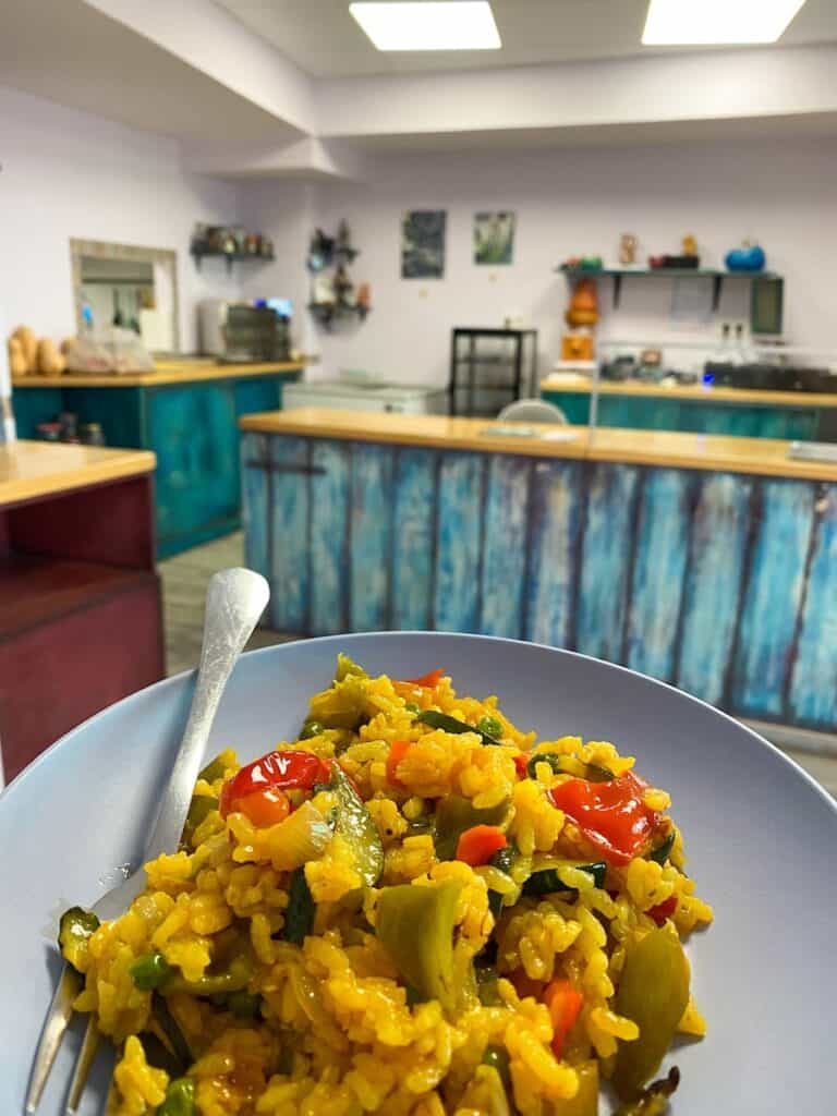 A close-up of a plate of golden, saffron-infused rice mixed with green and red vegetables, served in a cozy deli setting with turquoise-colored counters and rustic decor. The dish radiates warmth and freshness, capturing the inviting atmosphere of the eatery.