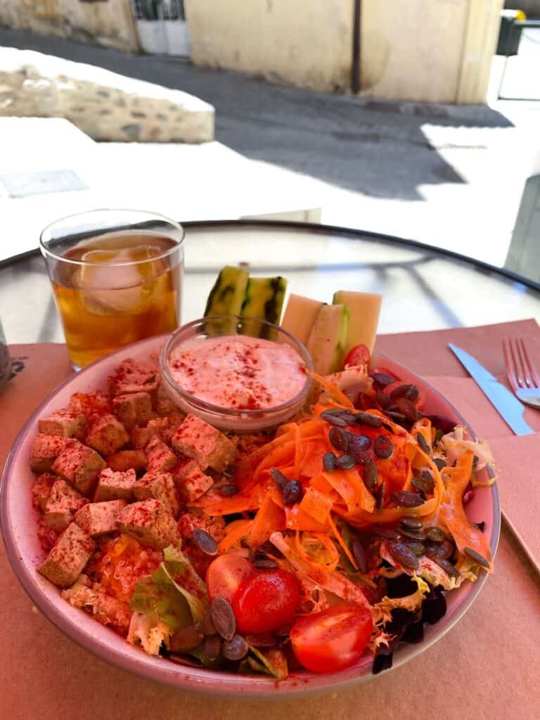 A vibrant bowl of salad topped with a mix of colorful veggies, including cherry tomatoes, shredded carrots, and crunchy seeds, alongside tofu cubes dusted with paprika. A glass of iced tea and a small dish of dipping sauce sit nearby, creating a refreshing meal setup in an outdoor café setting.