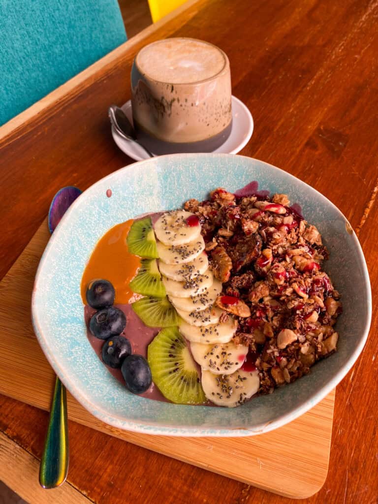 A colorful smoothie bowl topped with sliced banana, kiwi, blueberries, chia seeds, and a crunchy granola mix, arranged artfully for a fresh and balanced look. The bowl features a swirl of purple smoothie base with a spoonful of bright orange nut butter. In the background, there's a frothy cappuccino in a ceramic mug, adding a cozy touch to this vibrant breakfast scene.