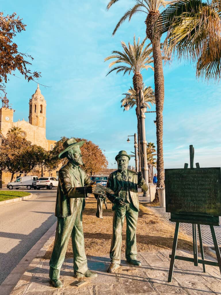 A statue of two artists and a canvas in front of the church in Sitges, you can see this on a day trip from Barcelona to Sitges by train