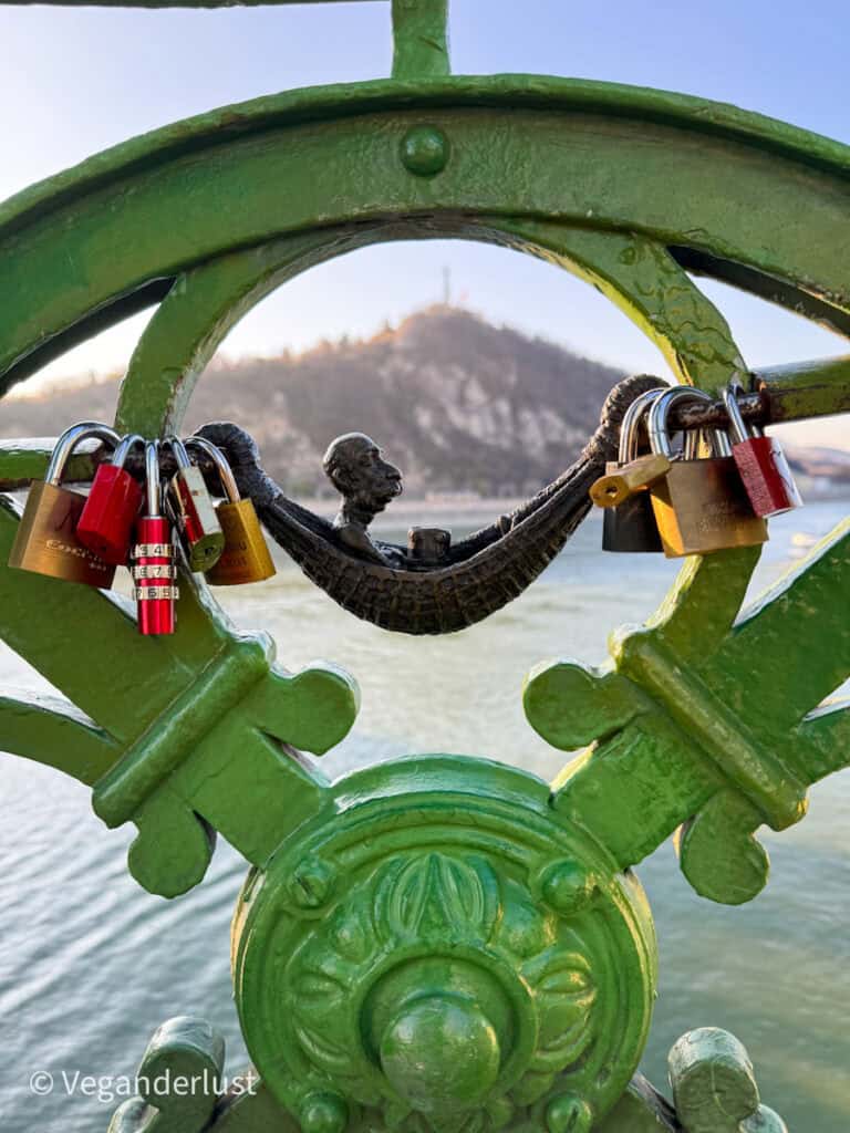 This image features a small, whimsical bronze statue of a reclining figure on Budapest’s Liberty Bridge, surrounded by colorful love locks attached to the green railing. In the background, Gellért Hill and the Danube River provide a scenic view, softly blurred to highlight the detailed foreground.