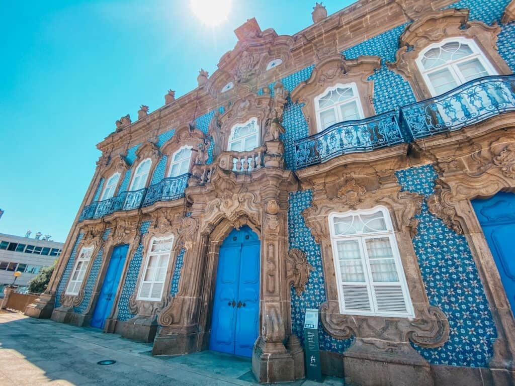 Bright blue doors and windows pop against an ornately detailed baroque façade adorned with traditional Portuguese azulejo tiles in a floral blue-and-white pattern. The building’s stone carvings and wrought iron balconies add elegance under a clear sky with the sun shining overhead.