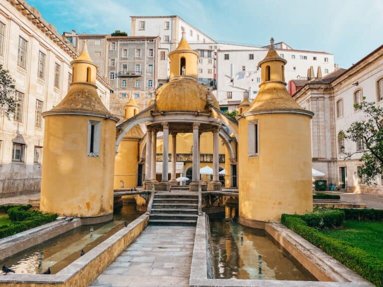 A striking yellow Baroque-style structure with domed towers and elegant arches sits in a courtyard, surrounded by reflecting pools and greenery. Small water channels run alongside, with pigeons perched near the edges. In the background, classic European buildings with white facades and shuttered windows rise against the blue sky.