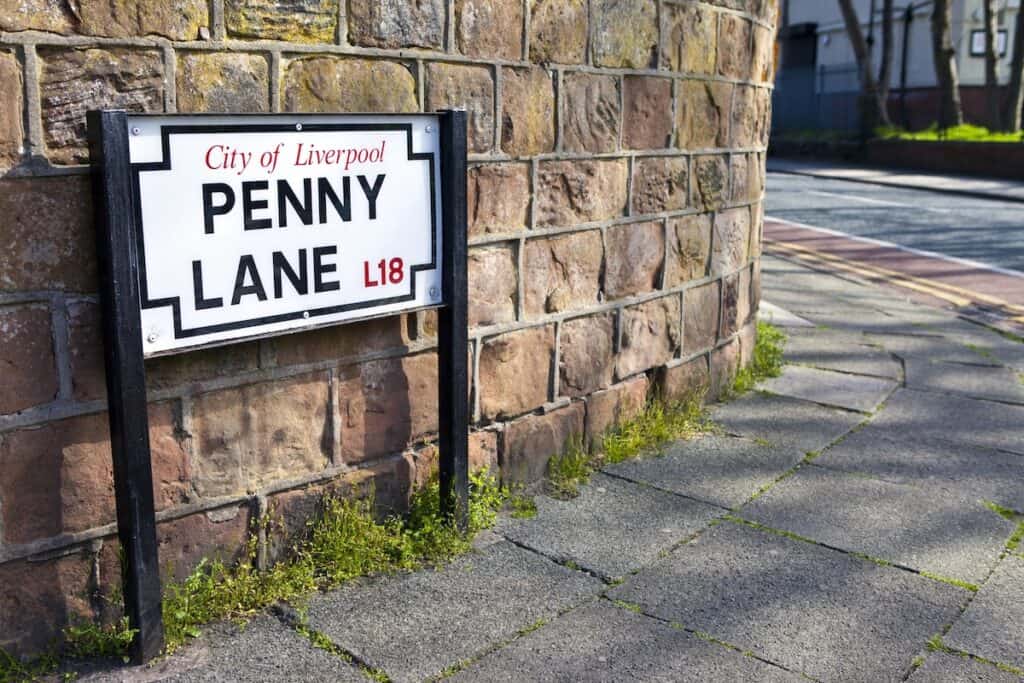 Street sign reading “City of Liverpool – Penny Lane L18” mounted on a brick wall at a curved sidewalk corner, referencing the iconic location made famous by The Beatles. The scene is quiet and well-lit by daylight, with stone pavement and a bit of greenery at the wall’s base.