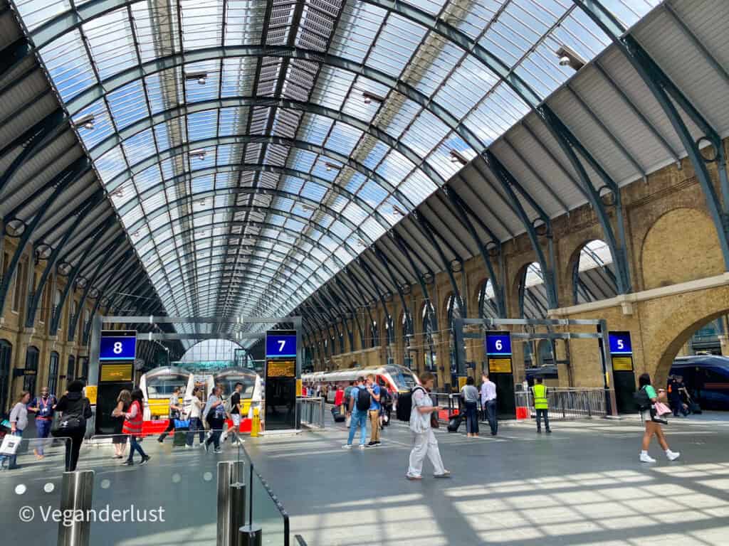 Interior of King's Cross station in London with a sweeping glass roof and iron beams above platforms 5 to 8 as travelers walk across the concourse and trains wait at the platforms. The airy design and visible platform signs emphasize the scale and activity of the station.