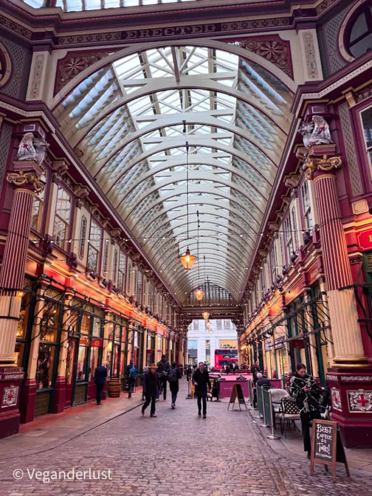 Covered Victorian arcade in London with a high glass ceiling and ornate red and cream columns lining a cobblestone walkway as people stroll between small shops and cafes. A hanging sign reading Erected spans the arch above and a chalkboard sign on the right reads Best coffee in town outside a cafe seating area.