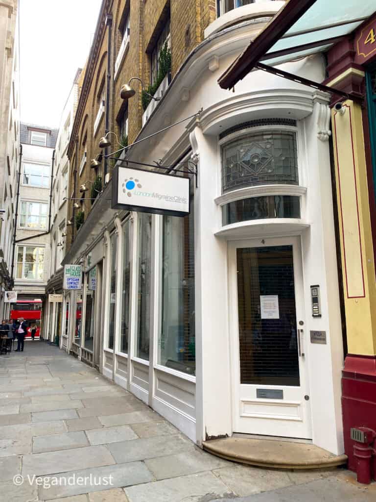 Narrow London street with a white doorway marked number 4 beneath a hanging sign that reads London Migraine Clinic and smaller text National Migraine Centre. The glass door has a printed notice and is set between shopfront windows and brick buildings along a quiet pedestrian lane.