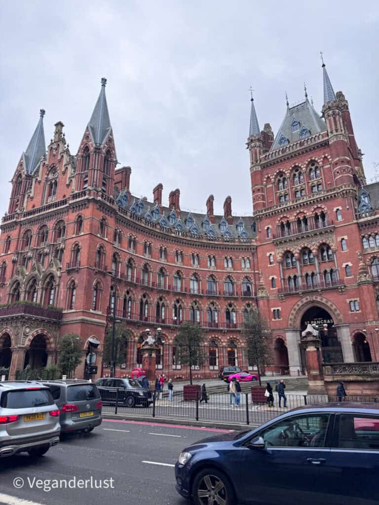 Exterior of the St Pancras Renaissance Hotel in London showing the red brick Gothic Revival facade with tall spires and arched windows as cars and pedestrians pass in front of the entrance marked St Pancras. The grand architecture highlights the historic character of the building near St Pancras station.