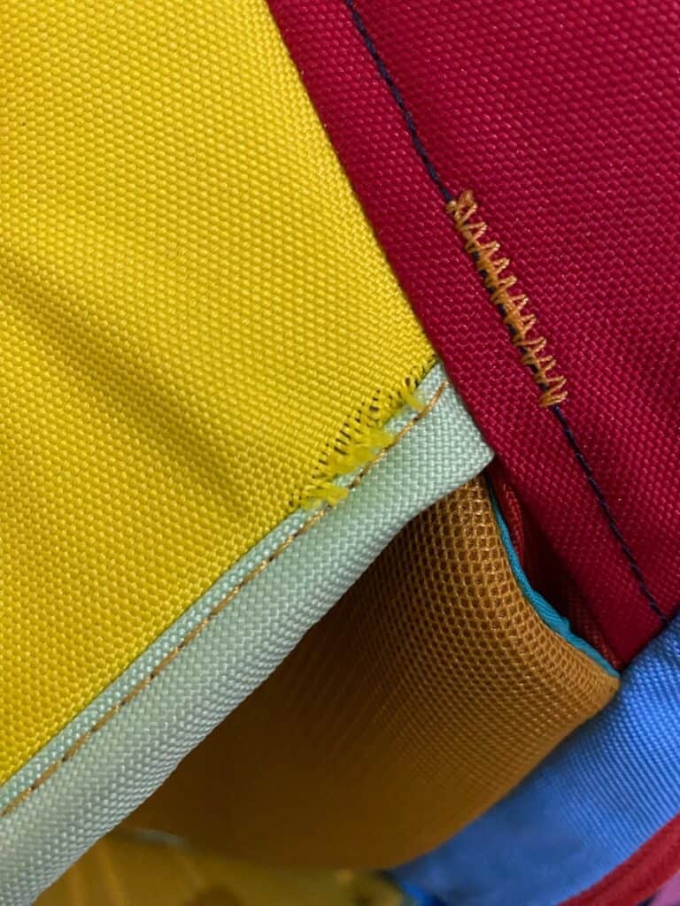 A close-up of a damaged section of a Cotopaxi backpack, showing frayed stitching and a small tear in the mustard-yellow fabric near a seam. The red, blue, and light green fabric sections are stitched together, with some uneven stitching visible.
