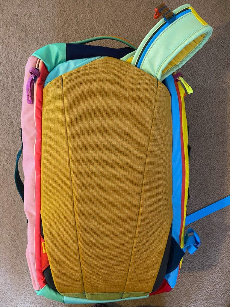 A colorful Cotopaxi backpack lying flat on a beige carpet, showing its mustard-yellow padded back panel. The pack features a mix of pastel and bright colors, including pink, teal, red, blue, and yellow, with multiple zippers and straps. One shoulder strap is partially visible at the top.