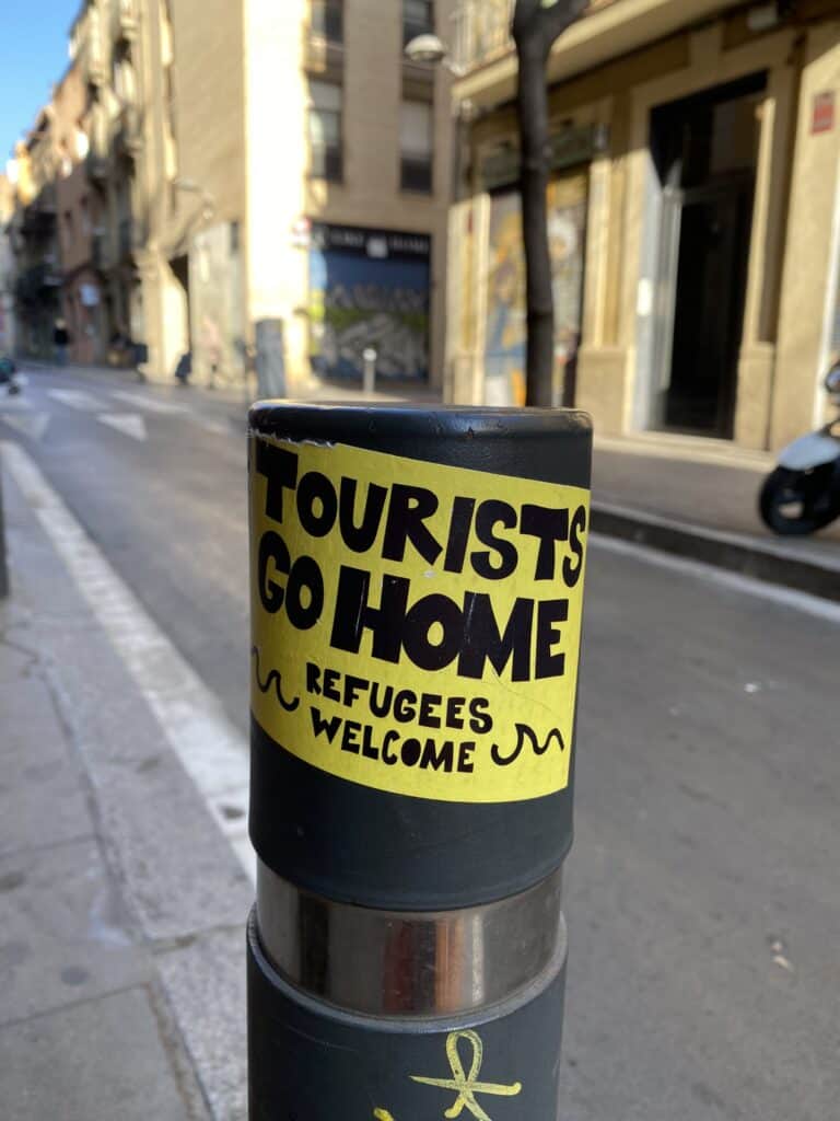 Bright yellow sticker on a street post reads "TOURISTS GO HOME. REFUGEES WELCOME." on a quiet city street in warm sunlight.