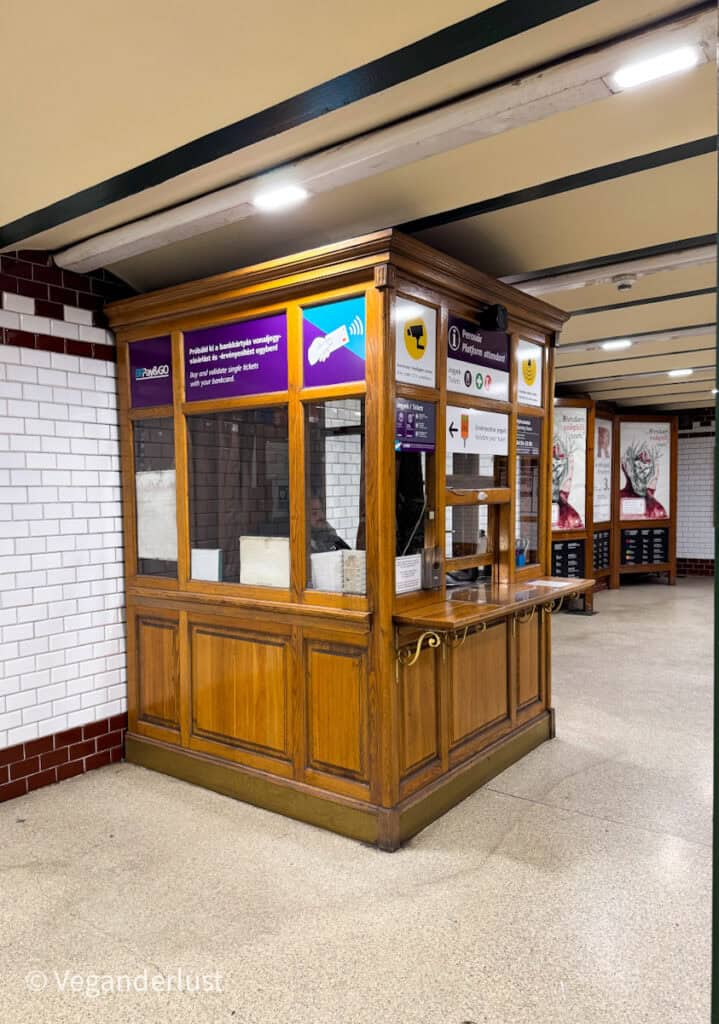This image depicts a vintage-style wooden ticket booth located inside a Budapest metro station. The booth is surrounded by tiled walls and features modern signage promoting contactless payment options alongside a classic design. The combination of old-world charm and contemporary functionality highlights the blend of history and modernization in public transport infrastructure.