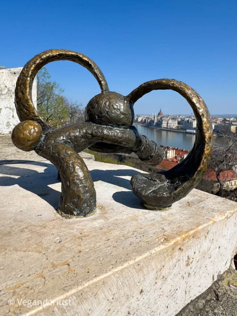 Bronze sculpture of a small stylized dog with looped ears mounted on a stone ledge, overlooking a wide river and a city skyline with a domed building in the distance under a clear blue sky. The foreground shows the textured metal surface of the statue while rooftops and the river stretch out behind it.