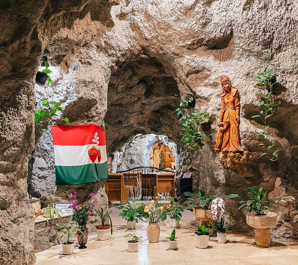 This image showcases a unique cave-like church interior with textured stone walls, a Hungarian flag prominently displayed, and various plants in decorative pots arranged on the tiled floor. A wooden statue of a saint is mounted on the rock wall, adding to the sacred atmosphere. The space blends natural elements with religious symbolism, creating a serene and spiritual ambiance.
