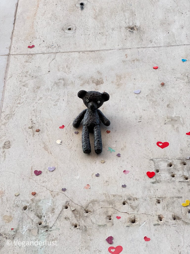 This image shows a small, dark-colored teddy bear statue mounted on a textured, weathered concrete wall. The wall is adorned with colorful, heart-shaped decorations, adding a whimsical and unexpected contrast to the urban setting. The artistic installation suggests a mix of street art and sentimentality.