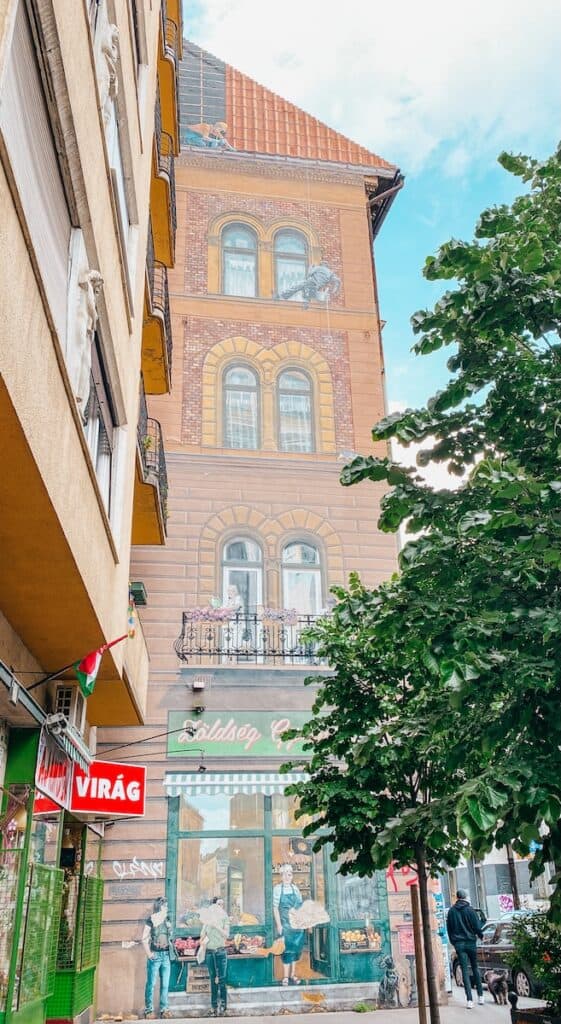 This photo shows a vibrant piece of street art on a Budapest building, blending seamlessly into the architecture. The artwork depicts a local grocery shop scene at ground level, complete with painted figures interacting near the storefront labeled "Zöldség Gyümölcs" (Vegetables & Fruits), and a balcony above with a figure and flowers, creating a whimsical, lifelike illusion. The surrounding urban environment includes trees and a shop sign reading "Virág" (Flowers).