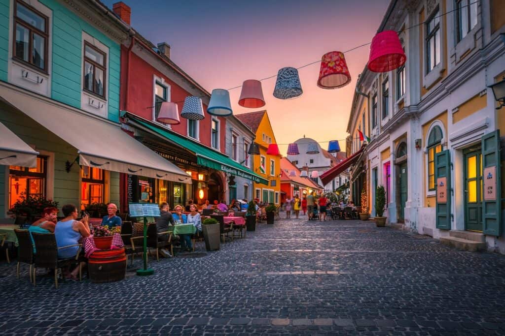 A colorful pedestrian street in Szentendre at sunset, lined with vibrant historic buildings, outdoor cafes, and hanging lampshade decorations strung across the cobblestone walkway. People are dining and strolling, creating a lively yet relaxed atmosphere under the warm glow of the evening sky.