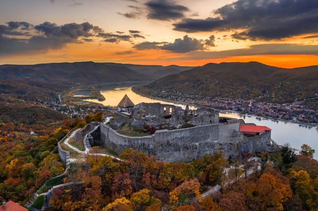A dramatic aerial view of Visegrád Castle overlooking the Danube River at sunset, surrounded by lush autumn foliage in shades of orange and yellow. The ancient stone fortress contrasts against the serene river and rolling hills, with scattered villages visible in the distance under a partly cloudy sky.