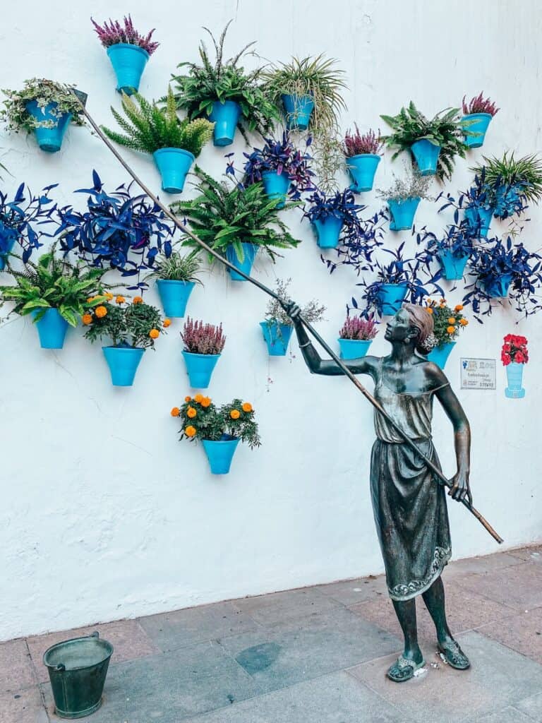 This image shows a decorative wall installation featuring bright blue flower pots filled with various vibrant plants, mounted in a cascading pattern on a whitewashed wall. In the foreground, a bronze statue of a woman holding a long-handled tool appears to interact with the pots, adding a lifelike element to the artistic display. A small metal bucket is placed on the ground near the statue, enhancing the scene's rustic charm.
