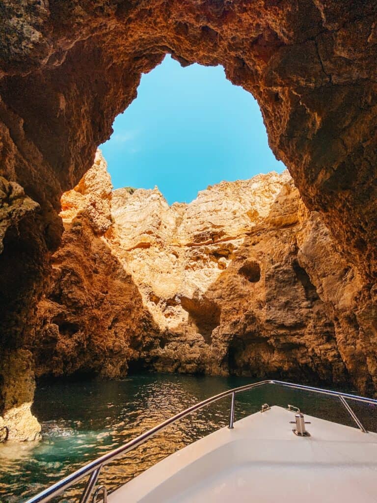 View from a boat entering a sea cave with rugged golden rock formations and turquoise water, framed by a jagged arch opening to a bright blue sky. Sunlight highlights the textures of the cave walls, creating a warm contrast against the cool shadows and water below.