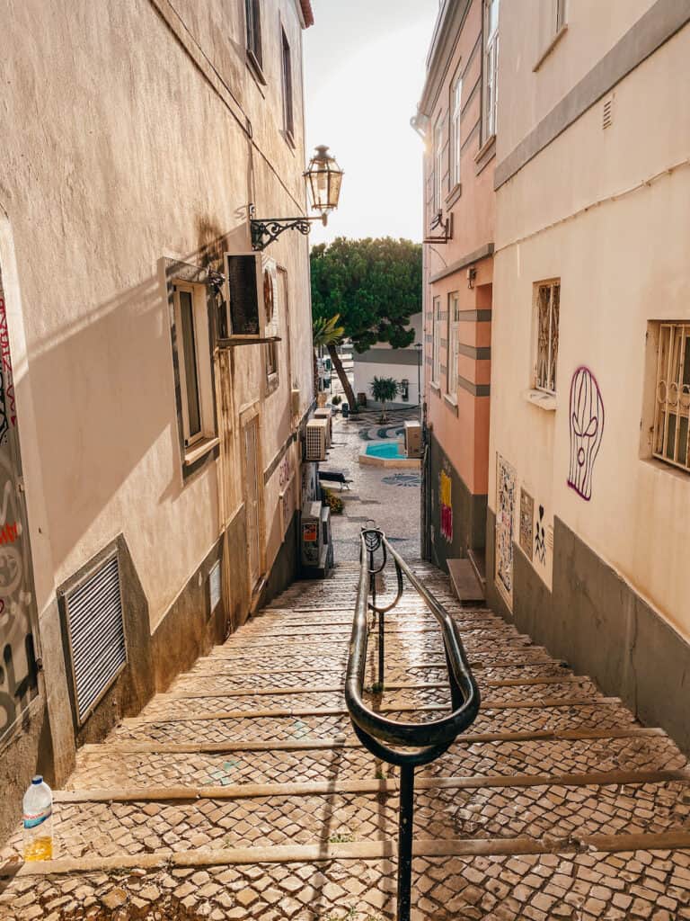 A narrow, sunlit alleyway in a European city features a steep stone staircase with a central iron handrail, lined by old buildings with graffiti and street art. At the bottom, a small courtyard with a tree and blue circular pattern is visible, adding charm and color to the quiet, urban scene.