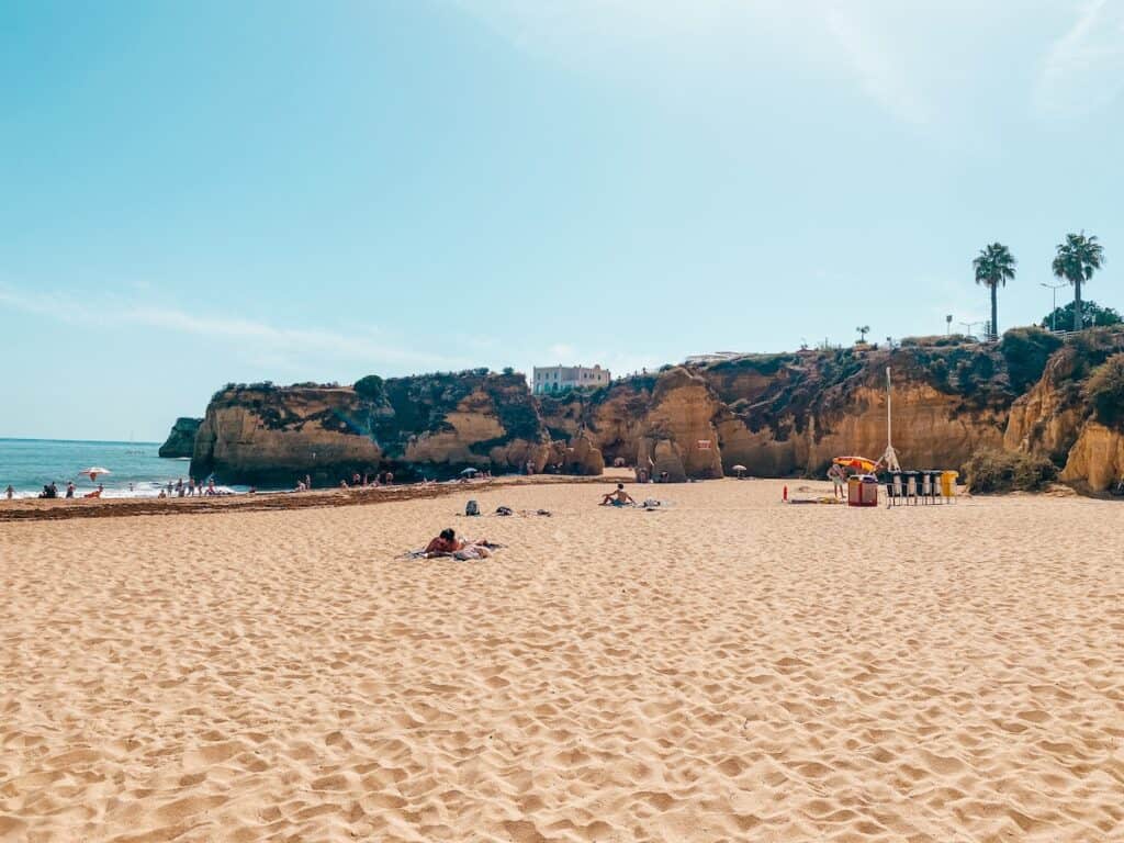 Wide sandy beach with scattered sunbathers, backed by golden cliffs and a few palm trees under a clear blue sky. A small cluster of colorful umbrellas and lifeguard equipment sits near the cliffside, while the calm sea and distant rock formations frame the left side.