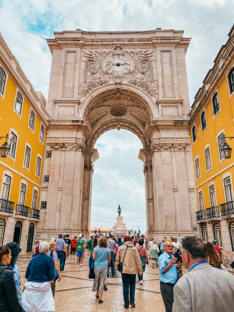 A big arch next to yellow buildings in Lisbon, in the back you can see a small statue straight behind the arch