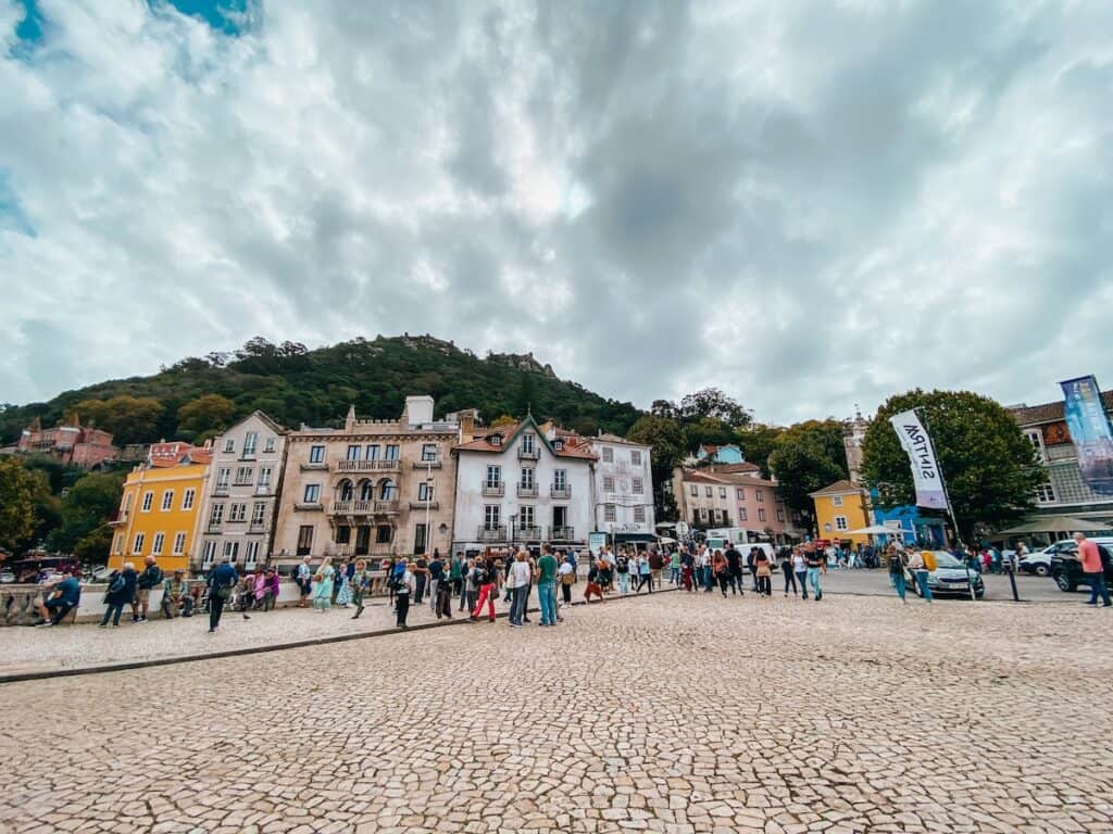 Crowd of tourists in a cobblestone town square in Sintra, Portugal, surrounded by colorful, historic buildings with forested hills in the background. The overcast sky looms above, and a white flag marked “Sintra” flutters near a group of parked cars and food trucks.