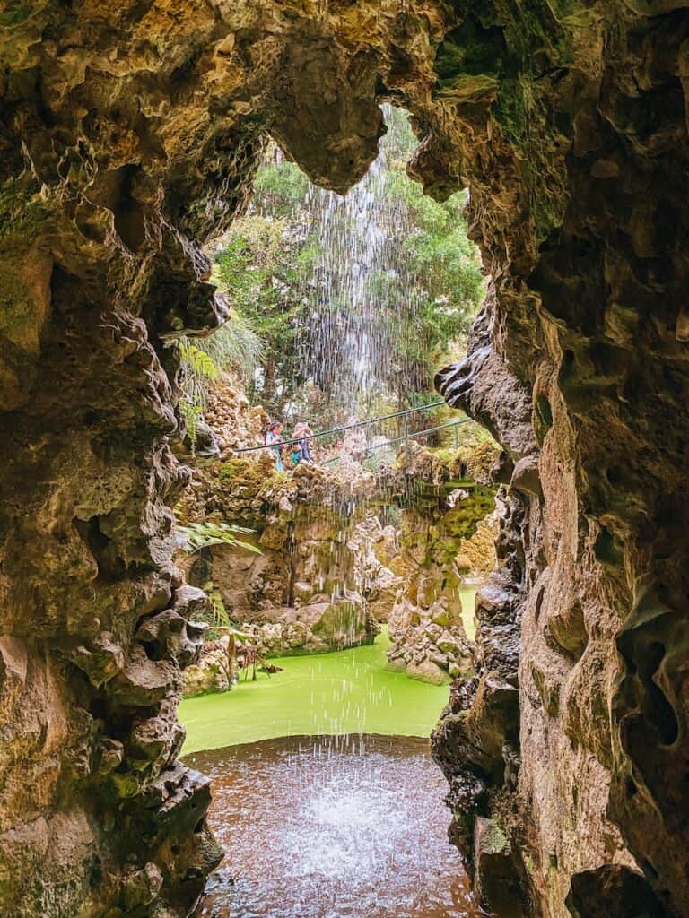 View from inside a rocky grotto looking out through a curtain of falling water into a lush garden with green water and stone pathways. Two people can be seen in the distance crossing a small bridge surrounded by greenery and rugged rock formations.