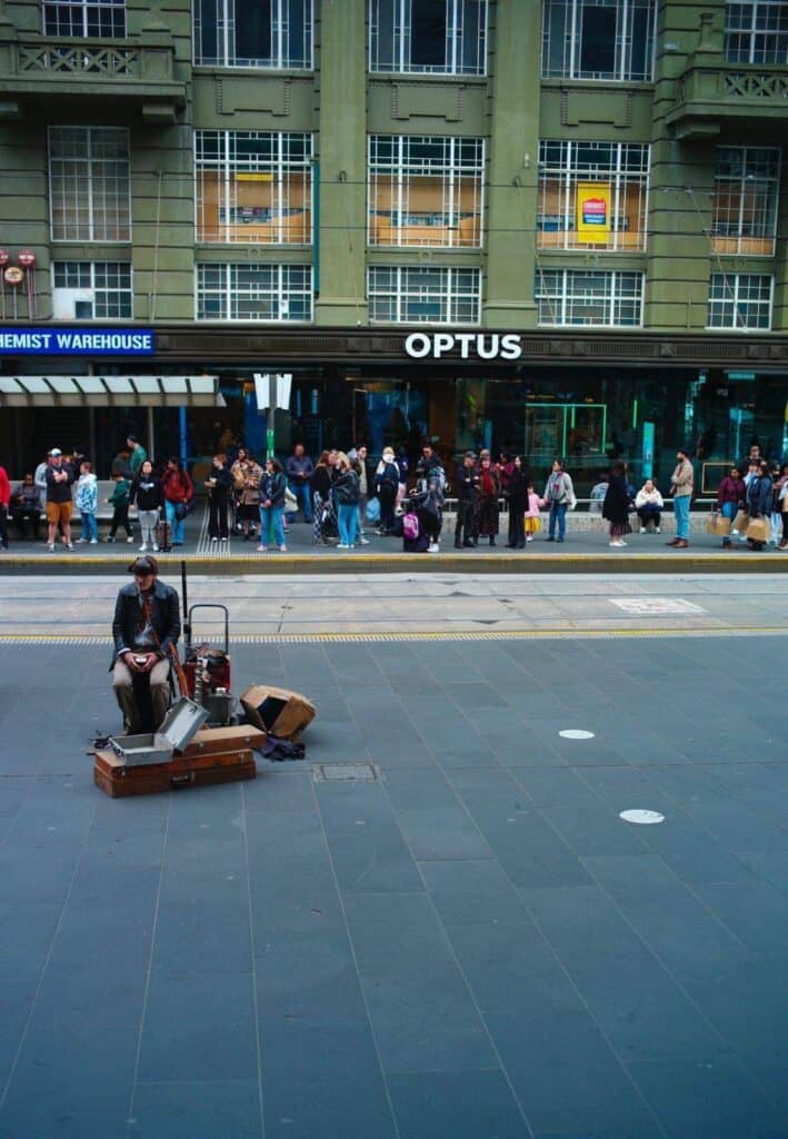 A guy playing an instrument on the street, in the background people are waiting for the tram
