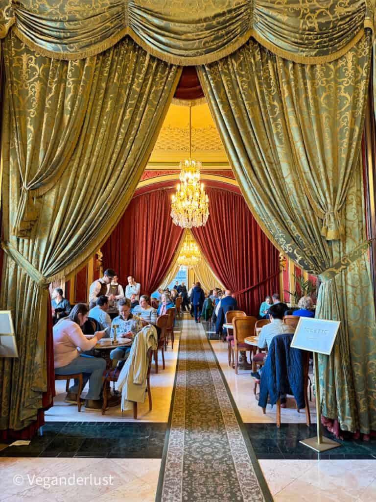Elegant café interior with tall green patterned curtains framing a long room filled with small tables where people sit and eat, under ornate chandeliers and red drapes, with a sign reading "TOILETTE" on the right side.