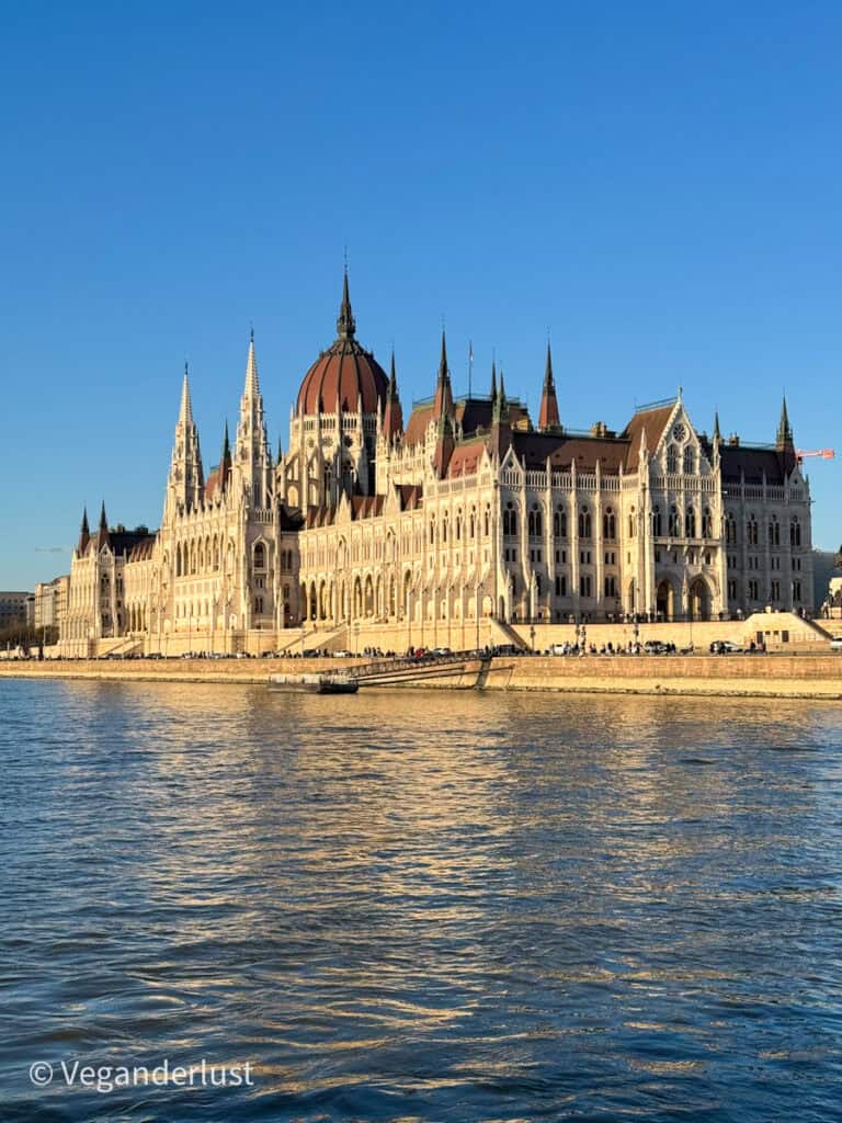 The Hungarian Parliament building along the Danube River under a clear blue sky, with its white facade, pointed spires, and large red dome reflected in the water as people walk along the riverbank.