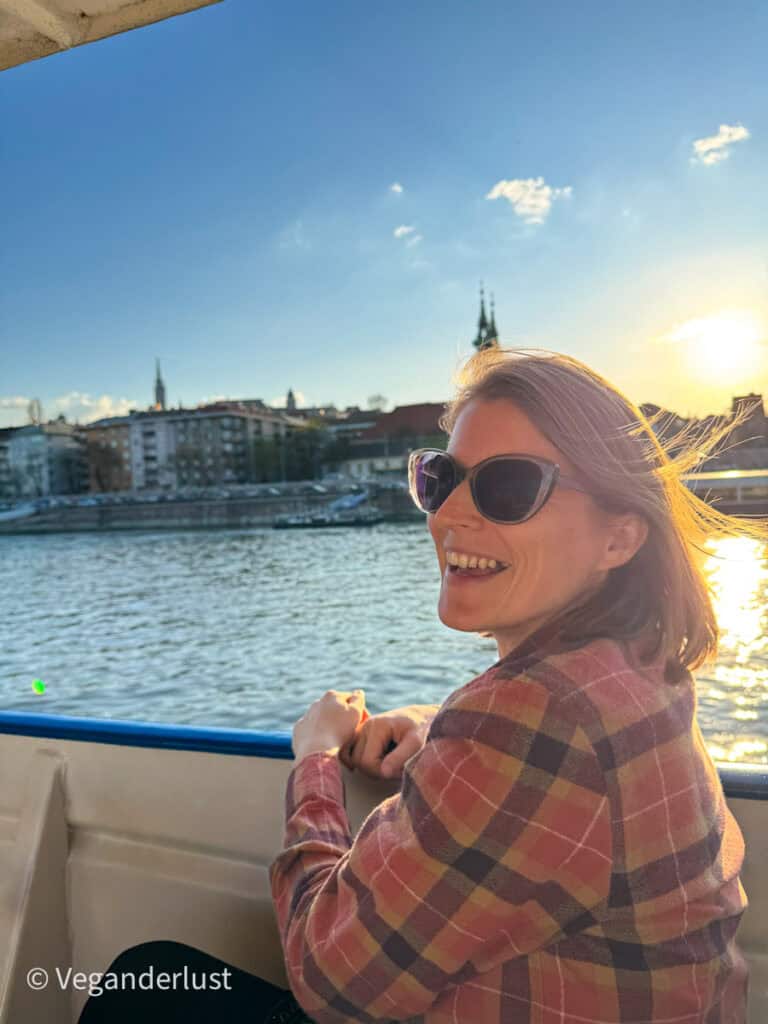 Tina wearing sunglasses and a plaid jacket smiling while leaning on the railing of a boat on the Danube River at sunset, with city buildings and church spires softly lit in the background.