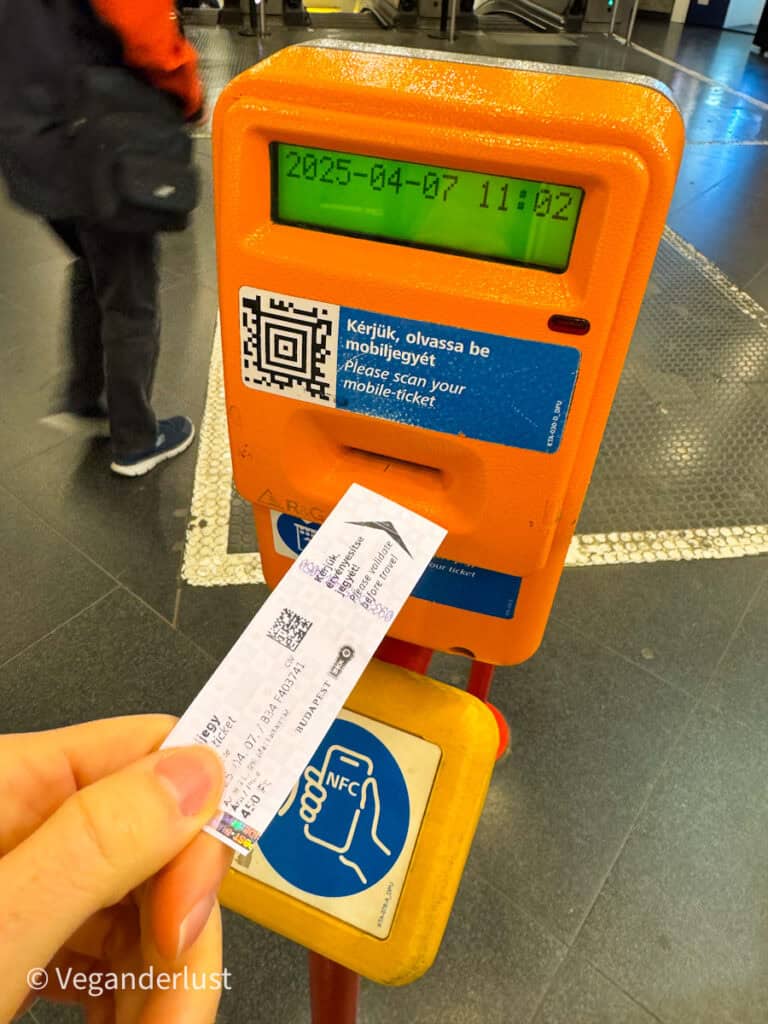 Hand holding a printed transit ticket up to an orange ticket validation machine in a subway station, with the screen displaying "2025-04-07 11:02". The machine shows a QR code and the text "Kérjük, olvassa be mobiljegyét" and "Please scan your mobile-ticket" alongside an NFC symbol, while blurred commuters walk past in the background.