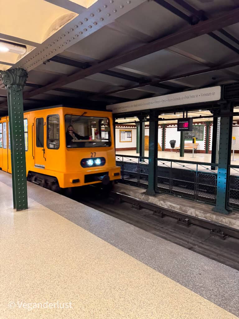 Bright yellow metro train pulling into an underground station with green metal columns and tiled walls, while a digital sign in the background displays "-30" and a platform sign lists stops including "Örs vezér tere" and "Deák Ferenc tér". The train approaches along the tracks with headlights on as passengers wait on the opposite platform.
