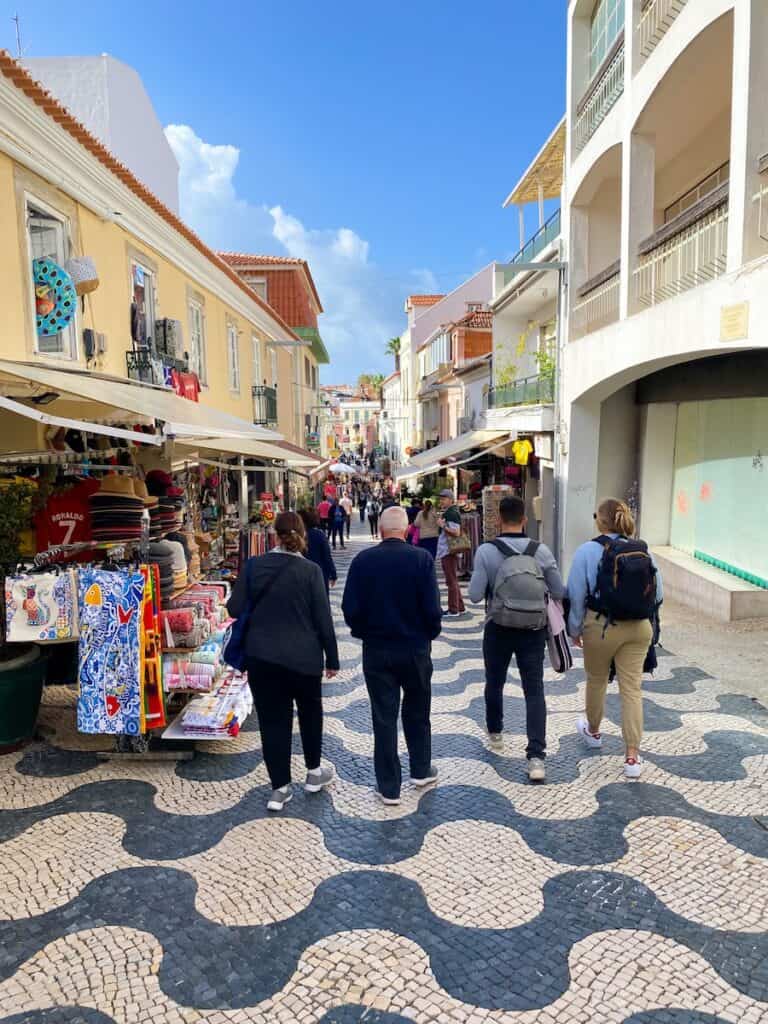 Bustling pedestrian street lined with colorful souvenir shops and cafes, featuring a distinctive black-and-white wave-patterned cobblestone walkway. Shoppers and tourists stroll under a bright blue sky, surrounded by pastel buildings with red-tiled roofs and open balconies.