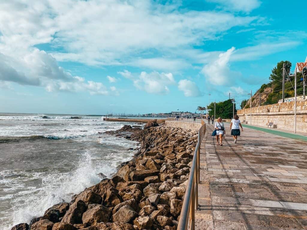 Stone-paved coastal promenade with two women walking alongside a rocky shoreline, as waves crash against the rocks. The path runs parallel to a stone wall with greenery and a tram line, under a sky filled with scattered clouds and bright blue patches.