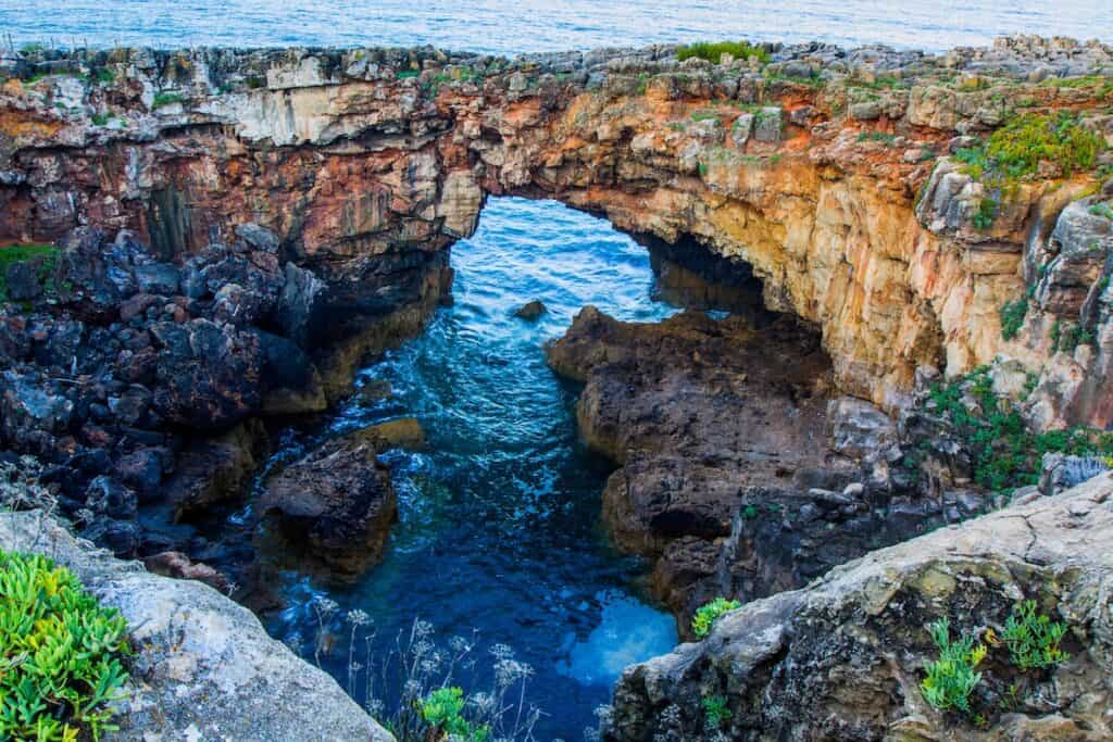 Natural sea arch carved into rugged coastal cliffs, with turquoise waves crashing into a rocky inlet below. The layered rock formations display shades of red, orange, and gray, surrounded by patches of green vegetation near the cliff’s edge.