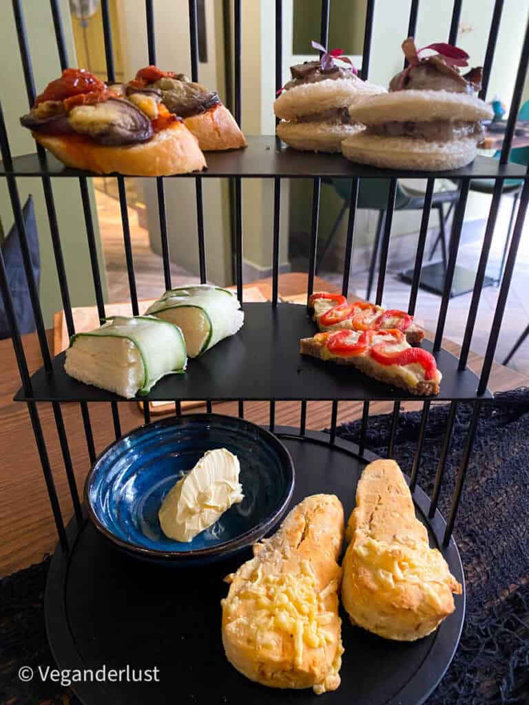 A tiered afternoon tea stand displays savory bites including sandwiches vegetable topped crostini and scones with cream. The black metal stand contrasts with the warm wooden table and green interior.