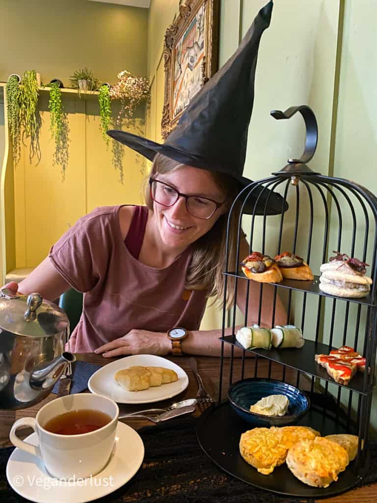 A woman in a witch hat pours tea from a silver teapot beside a tiered tray of savory snacks and scones. A white teacup and plate sit ready on the table in front of her.