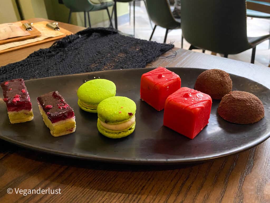 A black platter presents colorful mini desserts including bright green macarons red glazed cakes layered sponge slices and chocolate domes. The elegant sweets are neatly arranged for a modern afternoon tea experience.
