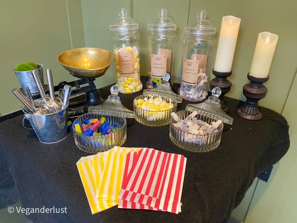 A Halloween themed candy station with glass jars labeled Sherbet Lemons Tongue Painters and Cauldron Cola Bottles filled with colorful sweets. Metal scoops striped paper bags vintage scales and flickering candles sit on a black table against green paneled walls.