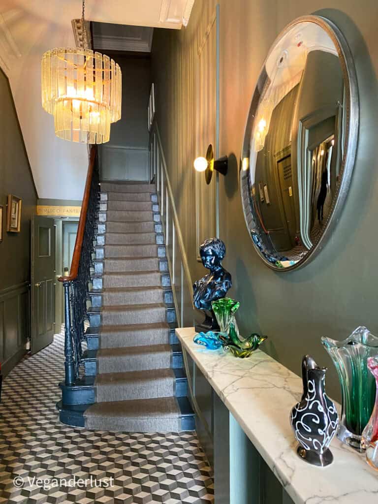 A narrow staircase with a beige runner leads upward beneath a glowing chandelier in a stylish hallway. A round wall mirror marble console table and decorative glass pieces add a boutique hotel feel.