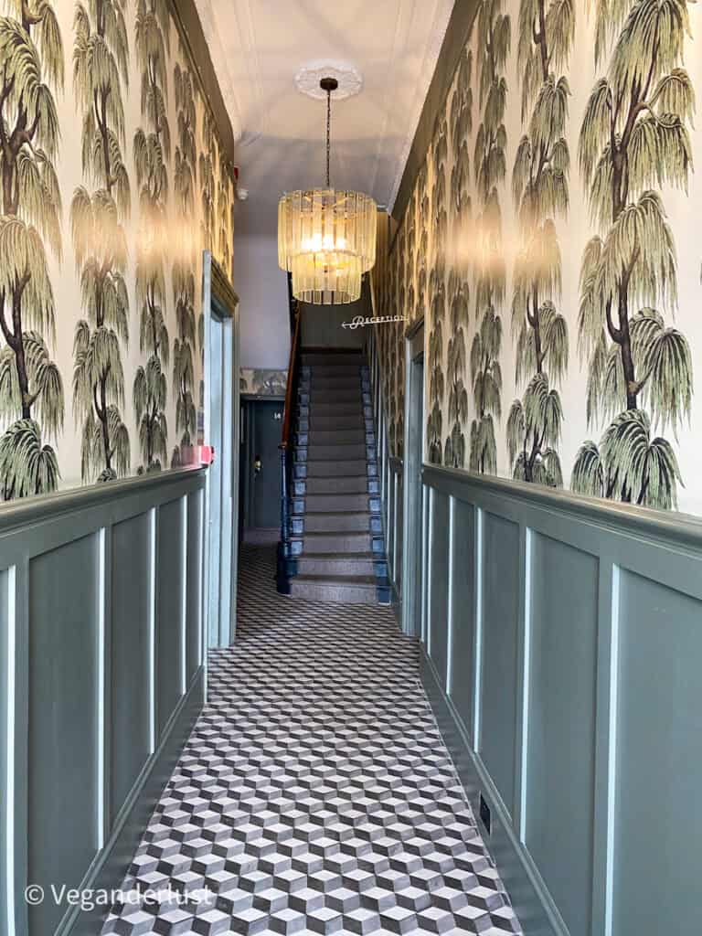 A long corridor with green paneling and palm tree wallpaper draws the eye toward a staircase at the end. Patterned tile flooring and a hanging chandelier create a dramatic and welcoming entrance.