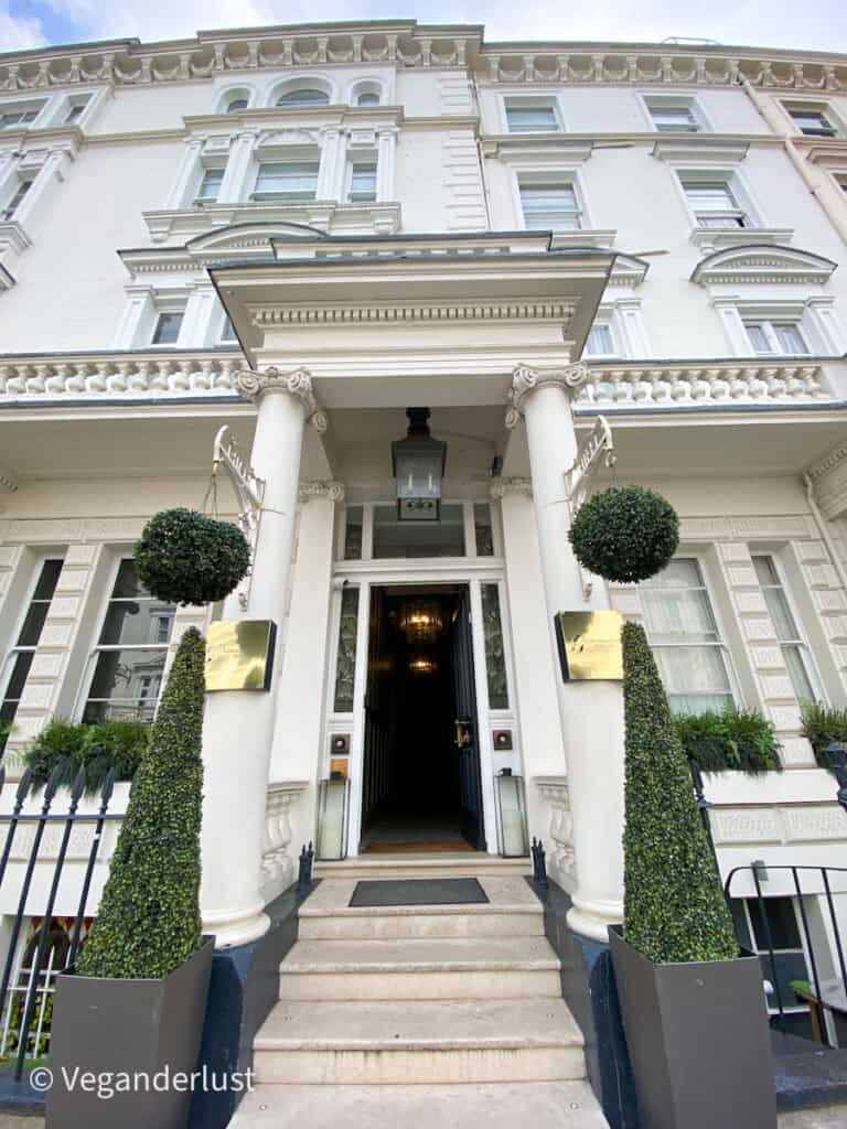 The exterior of a white townhouse hotel with tall columns and manicured topiary in planters framing the doorway. A gold sign and open front door invite guests inside.