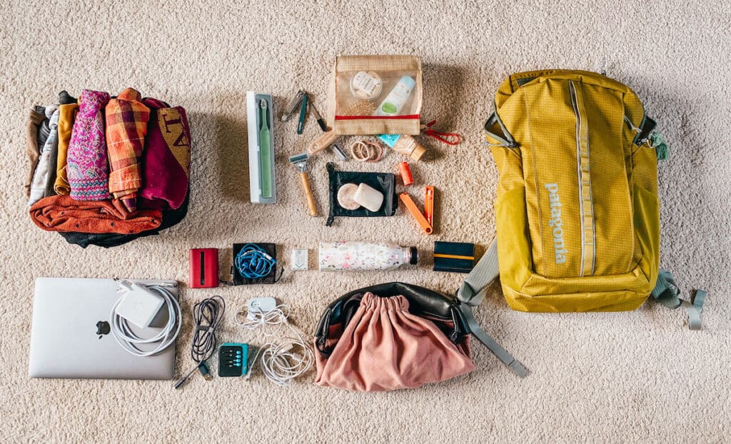 All items from a minimalist winter packing list displayed on a carpet, featuring clothing, electronics, a drawstring bag, a yellow backpack, toiletries, and travel documents.