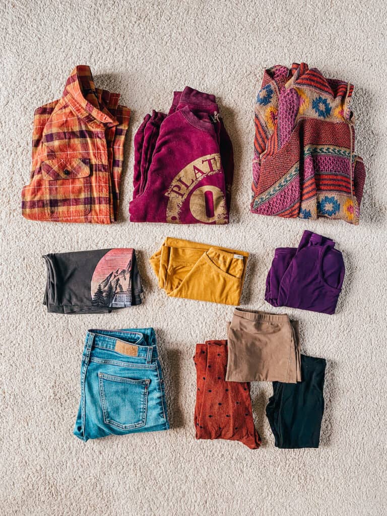 A variety of winter clothing neatly arranged on a carpet, including a plaid shirt, a purple hoodie, patterned sweaters, and assorted pants and shorts in earth tones. Everything you need for minimalist packing in winter