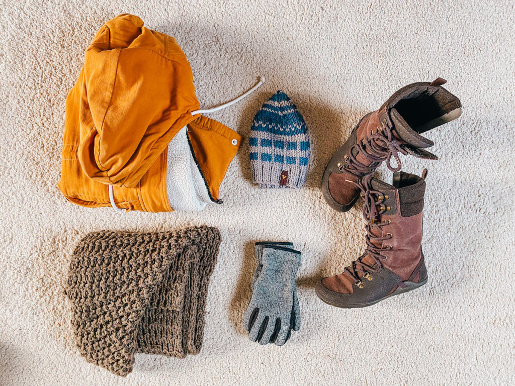 A mustard yellow winter jacket, a blue and white knitted beanie, a pair of brown lace-up winter boots, a chunky knit brown scarf, and a single grey glove displayed on a carpeted floor.
